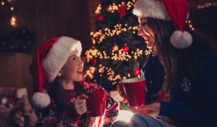 O fetita si mama ei stau langa bradul de Craciun si beau ciocolata calda